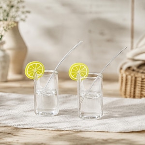 May include: Two clear glass shot glasses, each containing a thin, curved metal straw and a bright yellow-green lime slice garnish attached to the rim. The glasses are cylindrical with thick bases, filled with a clear liquid, and stand upright.