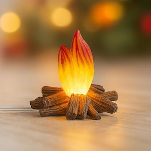 May include: A miniature campfire decoration with a glowing flame. The flame is a translucent yellow and orange, with red accents. The base is made of brown, textured logs. A small white wire extends from the base.