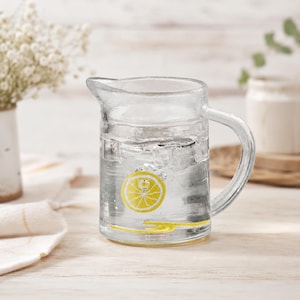 May include: A clear glass pitcher with a sturdy handle, filled with water and ice cubes. The exterior of the glass features a yellow lemon slice graphic on the front and a yellow lemon peel graphic near the base, adding a bright accent.