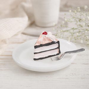 May include: A miniature slice of cake with light pink layers, dark brown frosting borders, and a dollop of white whipped cream topped with a single red cherry, resting on a small white plate next to a tiny silver-colored metal fork.