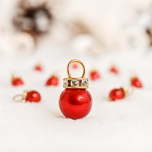 May include: A close-up of a red Christmas ornament with a gold loop and a band of clear crystals. The ornament is round and shiny, with a gold loop at the top. Other ornaments are blurred in the background.