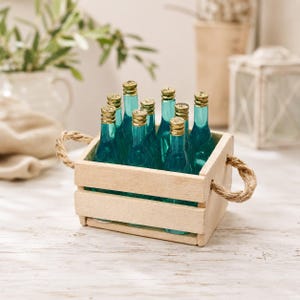 Könnte beinhalten: Eine kleine rechteckige Holzkiste mit zwei dicken Jute-Seilgriffen, die neun Miniatur-Glasflaschen enthält. Jede Flasche hat eine leuchtend blaugrüne Farbe mit einem metallisch goldenen Verschluss und steht aufrecht zwischen den Holzlatten.