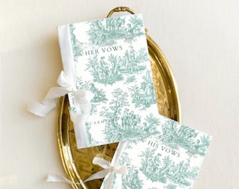 May include: Two wedding vow books with a teal and white toile pattern, tied with white ribbons, rest on a gold tray. The book covers read "HER VOWS" and "HIS VOWS". The books are labeled "CAROLINE & JOEY".