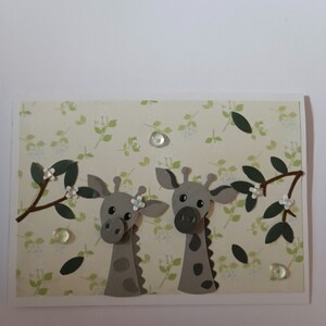Könnte beinhalten: Eine handgemachte Grußkarte mit einem grün-weißen Blumenmuster. Zwei graue Papiergiraffen mit weißen Blumen sind ausgeschnitten und auf die Karte gelegt.