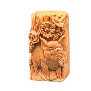 Può includere: Una saponetta rettangolare marrone con una scultura dettagliata di un maiale con fiori. Il maiale è rivolto verso il lato destro dell'immagine e ha un fiore in bocca.