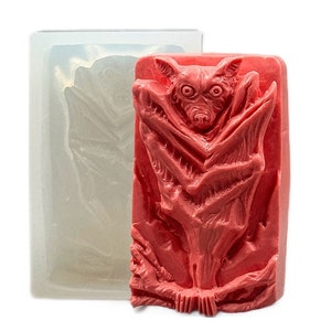 May include: A silicone mold for making a red bat-shaped soap or candle. The mold is made of clear silicone and has a detailed design of a bat with its wings spread out.