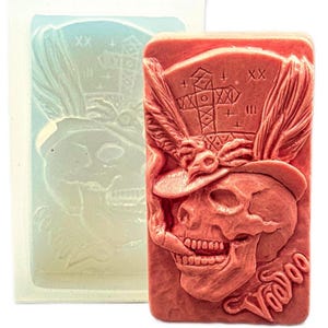 May include: A red rectangular soap bar with a skull wearing a hat and the word "Voodoo" carved into it. A clear mold of the same design is next to it. The skull has a detailed design with feathers and cross symbols.