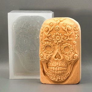 May include: A detailed sugar skull mold made of silicone. The mold is in the shape of a skull with intricate floral designs and a sugar skull design. The mold is light brown in color.