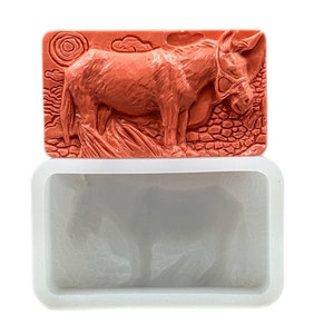 May include: A silicone mold for making soap or candles in the shape of a donkey. The mold is white and has a raised design of a donkey with a halter. The donkey is standing on a textured surface.