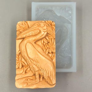 Puede incluir: Un pelícano marrón tallado en relieve, con plumas detalladas y un pico largo, sobre un fondo de follaje estilizado. La talla está en un molde rectangular, con un molde de silicona a juego al lado.
