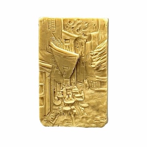 Könnte beinhalten: Eine goldfarbene Seife mit einem Reliefdesign einer europäischen Straßenszene. Die Szene zeigt Gebäude, ein Café und Menschen, die spazieren gehen.