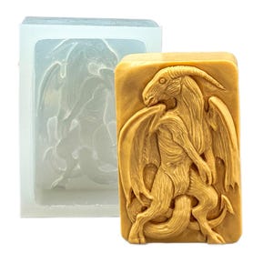 May include: A tan-colored bar of soap with a detailed dragon carving, next to a clear silicone mold of the same design. The dragon has horns, wings, and a serpentine tail. The soap is rectangular and stands out against the white background.