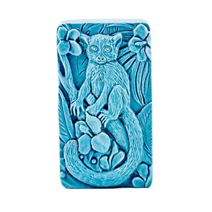 May include: A rectangular, turquoise-colored decorative soap bar featuring a detailed relief carving of a lemur sitting on a branch. The lemur is surrounded by leaves and flowers, creating a nature-themed design.