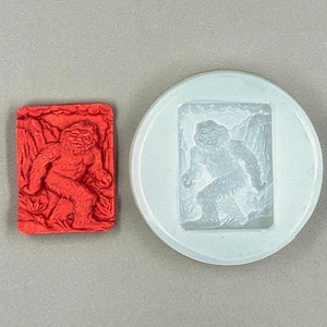 May include: A white silicone mould with a raised design of a Bigfoot walking through a forest. The mould is shown next to a red silicone casting of the same design.