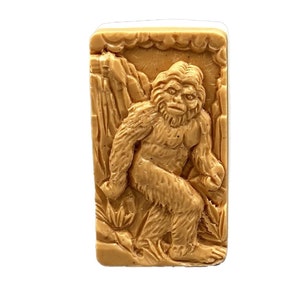 Puede incluir: Una pastilla de jabón marrón con una imagen en relieve de una criatura parecida a Bigfoot de pie en un paisaje rocoso y montañoso.