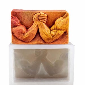 Può includere: Una saponetta rettangolare, color arancione, con un design scolpito di braccio di ferro. Sotto, è visibile uno stampo in silicone bianco con lo stesso motivo di braccio di ferro. La saponetta ha una superficie strutturata e dettagli muscolari.