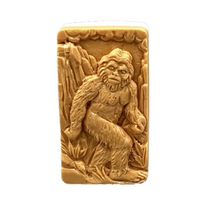 May include: A brown soap bar with a raised image of a Bigfoot standing in a forest. The soap bar is rectangular and has a textured surface.