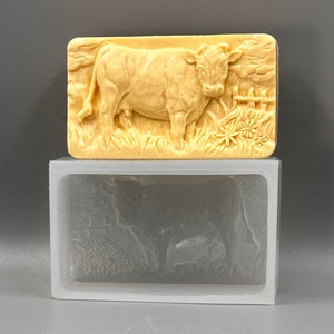 Peut inclure: Un moule en silicone beige avec un design détaillé d'une vache debout dans un champ d'herbe. Le moule est dans un récipient en plastique transparent.