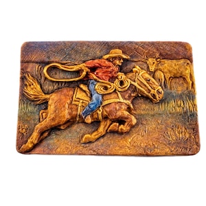 May include: A rectangular relief sculpture depicting a cowboy on horseback roping a steer. The cowboy wears a red shirt, blue jeans, and a brown hat. The horse is brown, and the background is a mix of brown and green.