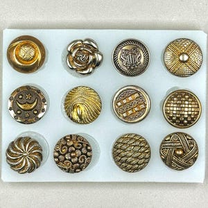 Op de afbeelding: Een wit dienblad met 12 gouden en zilveren knopen met verschillende ontwerpen. De knopen zijn in een rasterpatroon gerangschikt.