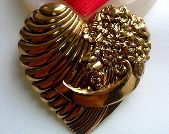 May include: A silicone mould in the shape of a heart with a raised design of a heart with flowers and a ribbon. The mould is on a white surface. Next to it is a gold-coloured heart-shaped brooch with a similar design.