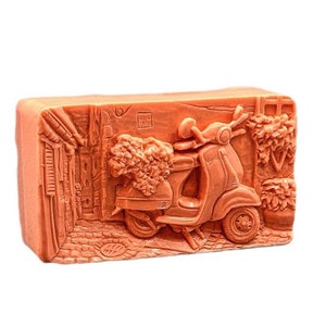Puede incluir: Una pastilla de jabón rectangular de color naranja con un diseño en relieve de una scooter conduciendo por una calle adoquinada con edificios a ambos lados.