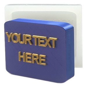 Può includere: Un cartello blu stampato in 3D con angoli arrotondati e il testo "YOUR TEXT HERE" in lettere dorate.