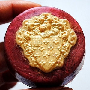 Op de afbeelding: Een ronde, rode en gouden decoratieve zeep met een reliëf, gouden, sierlijk ontwerp van een wapenschild.
