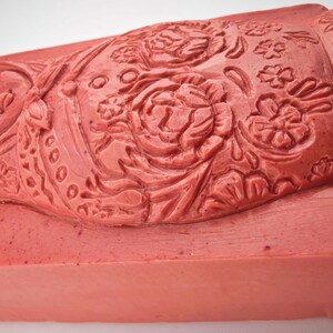 May include: A rectangular, coral-colored stamp with intricate floral and figure carvings. The design features a woman's face, roses, and other floral elements. The stamp is likely used for decorative purposes.