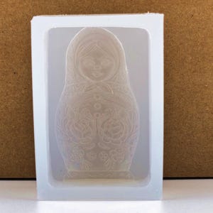 May include: A clear silicone mold in a rectangular frame, featuring a detailed relief of a Matryoshka doll. The doll design includes a headscarf, floral patterns, and button details. The mold is set against a neutral background.