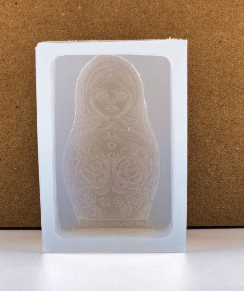 May include: A clear silicone mold in a rectangular frame, featuring a detailed relief of a Matryoshka doll. The doll design includes a headscarf, floral patterns, and button details. The mold is set against a neutral background.