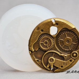 May include: A silicone mould for making jewellery or other crafts. The mould is shaped like a round gear with intricate details. The mould is white and the gear is a brass colour.