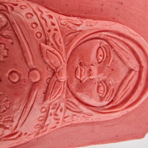 May include: A close-up of a red-toned carving of a Matryoshka doll. The doll's face and floral details are intricately carved. The carving is on a rectangular base.