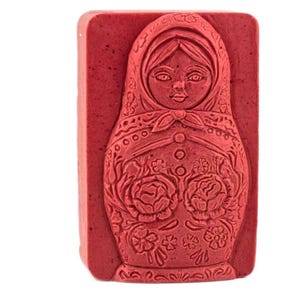 May include: A rectangular, red soap bar featuring a detailed relief of a Matryoshka doll. The doll's face, scarf, and floral patterns are intricately carved into the soap's surface. The soap is a solid, uniform color.
