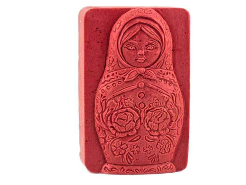 May include: A rectangular, red soap bar featuring a detailed relief of a Matryoshka doll. The doll's face, scarf, and floral patterns are intricately carved into the soap's surface. The soap is a solid, uniform color.