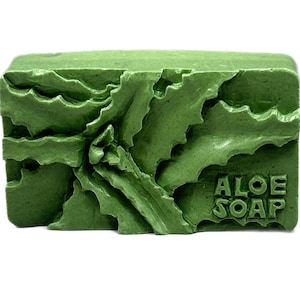 Könnte beinhalten: Eine grüne Seife in Form eines Blocks mit einer strukturierten Oberfläche und den eingeprägten Worten "ALOE SOAP" an der Seite.