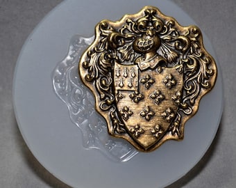 Pode incluir: Um emblema de latão detalhado, de estilo antigo, com um design de brasão, apresentando flor-de-lis e outros detalhes ornamentados. O emblema está sobre uma base circular cinza claro. O latão tem um acabamento quente e envelhecido.