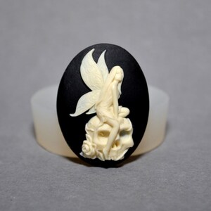 May include: A white cameo featuring a fairy sitting on a skull with a rose. The cameo is set in a black oval-shaped frame.