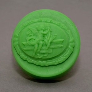 Può includere: Un sapone rotondo verde con un designo ovale in rilievo che presenta due cherubini che si abbracciano. L'ovale è circondato da un bordo decorativo.