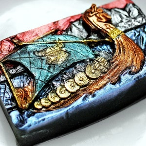 Puede incluir: Una escultura detallada de resina pintada a mano de un barco vikingo con un acabado dorado y cobre. El barco tiene una vela azul y verde con un diseño dorado y está decorado con monedas de oro.