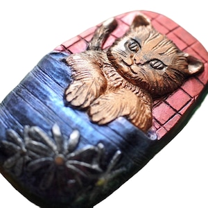 May include: A ceramic figurine of a brown cat sleeping on a red and blue patterned surface. The cat has a white chest and a white spot on its forehead.