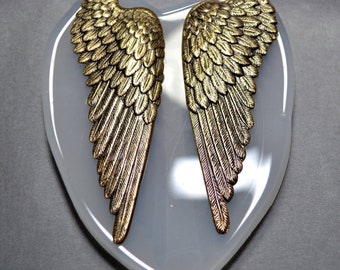 May include: Two bronze-colored angel wings are displayed on a heart-shaped, light gray surface. The wings have detailed feather textures and are positioned symmetrically. The background is a solid, medium gray.