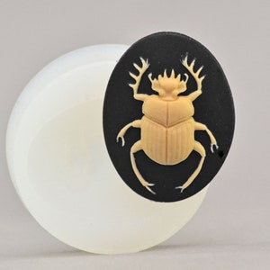 May include: A silicone mould for making resin jewellery featuring a detailed design of a beige scarab beetle on a black background.