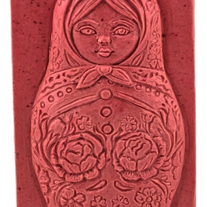May include: A rectangular, red soap bar featuring a detailed relief of a Matryoshka doll. The doll is adorned with floral patterns, a headscarf, and a button-down dress. The soap has a rustic, handmade appearance.