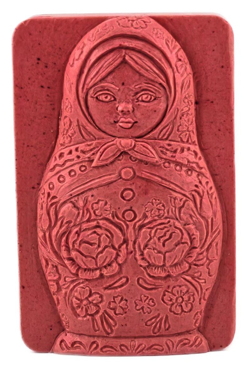 May include: A rectangular, red soap bar featuring a detailed relief of a Matryoshka doll. The doll is adorned with floral patterns, a headscarf, and a button-down dress. The soap has a rustic, handmade appearance.