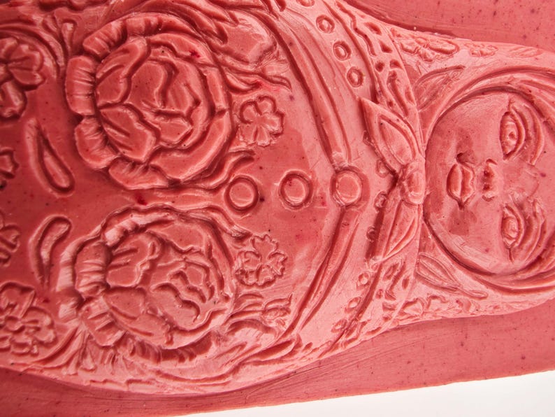 May include: Close-up of a detailed, coral-colored carving. The design features floral patterns, including roses, and a stylized face. The carving has a textured surface and is likely part of a larger decorative piece.