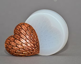 May include: A copper heart-shaped mould with a feathery design, next to a white silicone mould with the same design.