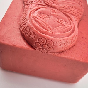 May include: A rectangular, coral-colored block with a carved relief of a woman's face and floral patterns. The detailed carving features a woman's face with closed eyes, surrounded by ornate floral and leaf designs. The block's edges are slightly rounded.