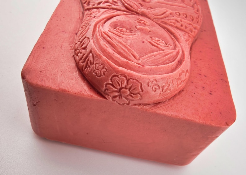 May include: A rectangular, coral-colored block with a carved relief of a woman's face and floral patterns. The detailed carving features a woman's face with closed eyes, surrounded by ornate floral and leaf designs. The block's edges are slightly rounded.