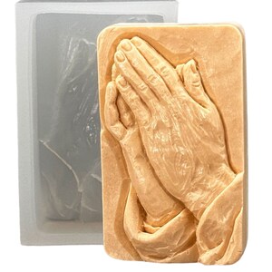 May include: A silicone mold for making soap or other crafts. The mold is shaped like two hands clasped in prayer. The mold is light brown in color and has a smooth, detailed surface.